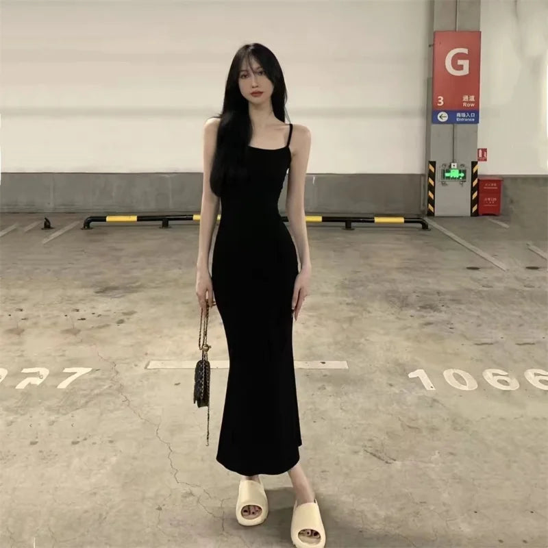 A woman in an Autumn Pure Sleeveless Dress Women Sexy Sheath Hotsweet Style Fashion All-match Vestido Feminino New Arrival Popular Chic by Maramalive™, stylish black midi dress and beige slide sandals stands in a parking garage holding a small purse, exuding an understated Korean style.