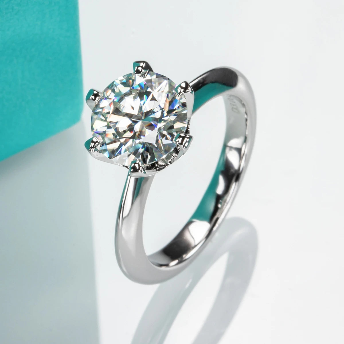 A luxurious silver ring with a large round-cut moissanite center stone in a six-prong setting, placed near a turquoise box. Introducing Moissanite Solitaire Rings: Stunning Engagement Jewelry by Maramalive™.