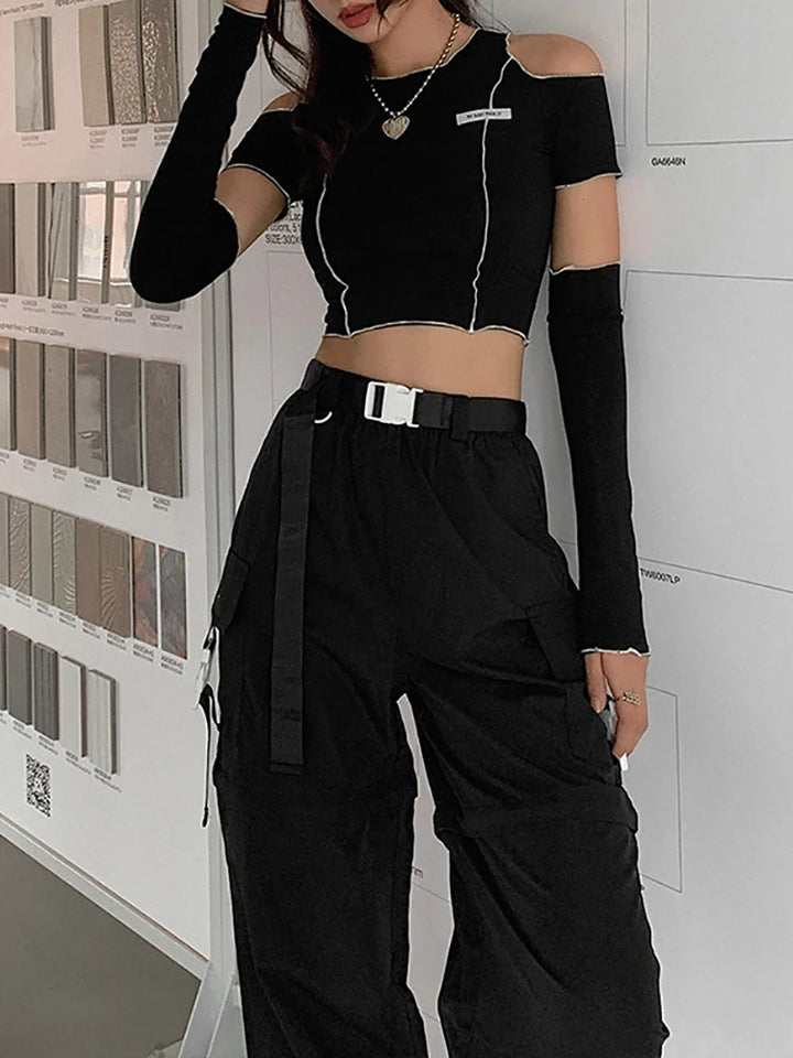 Person wearing a Goth Dark E-girl Style Patchwork Black T-shirts Gothic Open Shoulder Sleeve Y2k Crop Tops Ruffles Hem Hip Hop Techwear Women Tee from Maramalive™, black cargo pants, and a belt. They embody Gothic casual streetwear, standing in front of a wall with various samples or materials displayed.
