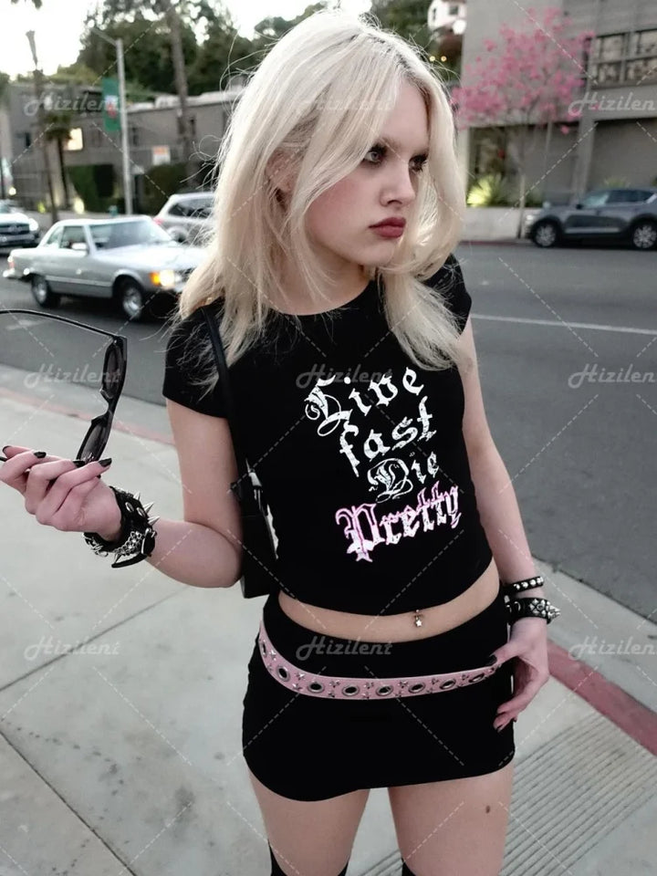 A woman with blond hair is standing on a sidewalk, holding sunglasses in one hand. She is wearing a black Maramalive™ 1Y2K clothes 2000s Streetwear Goth Short Sleeve T-shirts Fashion Letter Cute Baby Tees Summer E-girl EMO Crop Tops Slim and a black skirt with a pink belt.