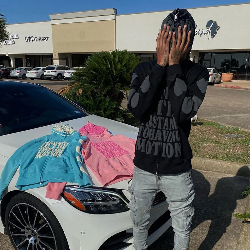 A person stands in front of a white car, covering their face with their hands. On the car’s hood are three Maramalive™ Goth letter printing Hoodies Y2K Zip Up Hoodie Women High Street Retro Hip Hop Casual Sweatshirt Fashion Hoodies in black, pink, and blue. A bustling High Street strip mall is visible in the background.