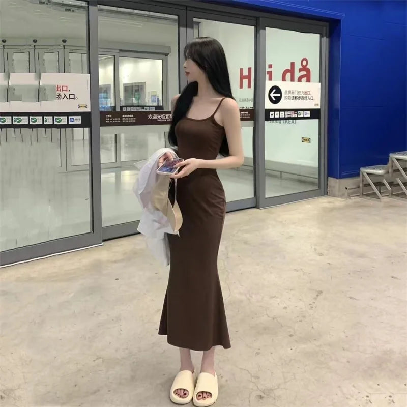 A woman in a long brown midi dress and white sandals, holding a phone and clothing, stands in front of a glass entrance with signs in Chinese. She is wearing the Maramalive™ Autumn Pure Sleeveless Dress Women Sexy Sheath Hotsweet Style Fashion All-match Vestido Feminino New Arrival Popular Chic.