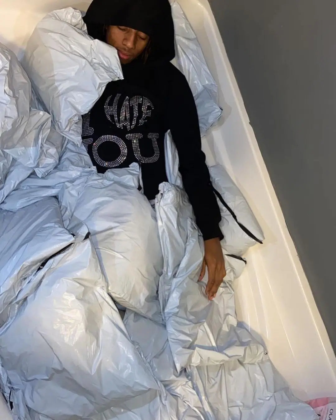 A person wearing a Maramalive™ Oversized Diamond skull Print Streetwear Hoodie Vintage Streetwear Tops Sweatshirt Goth Hoodies Women Y2k Clothes, showcasing High Street Style, is lying in a bathtub filled with plastic packages.