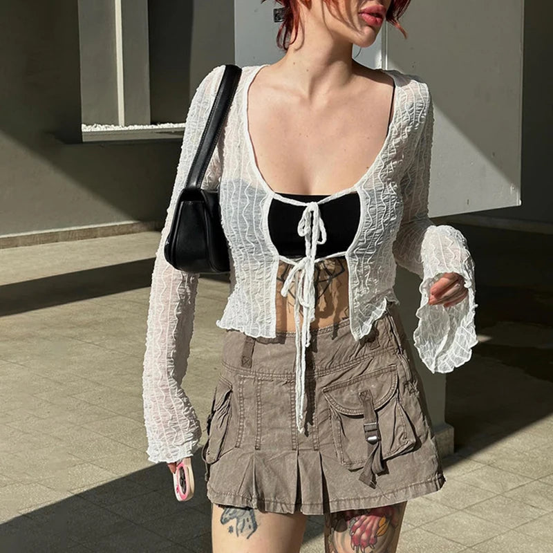 Person wearing a sheer white long-sleeve shirt over a black top, brown cargo mini skirt, and carrying a black bag. The Maramalive™ Goth Dark Folds Mall Gothic Lace Up Sexy Blouses Grunge Y2k Fairycore Long Sleeve T-shirts Female Transparent Skinny Fashion Tops adds a touch of Gothic fashion as they stand in a sunlit concrete area.