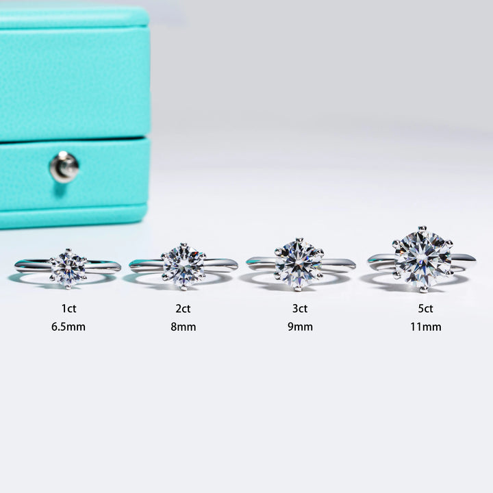 Four silver rings, each featuring a sparkling round-cut diamond (1ct, 2ct, 3ct, 5ct), are displayed next to a light blue box. The diamonds' sizes are labeled 6.5mm, 8mm, 9mm, and 11mm respectively. Consider Maramalive's Moissanite Solitaire Rings: Stunning Engagement Jewelry with a moissanite center stone for an equally stunning alternative.