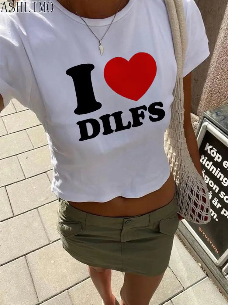 A person wearing a white 2000s I Love Dilfs Women T-shirt Summer Clothing women Crop Tops Slim Goth Short Sleeve letter T-shirts Y2K Fashion from the Maramalive™ brand and a green skirt, holding a mesh bag, stands on a paved path.