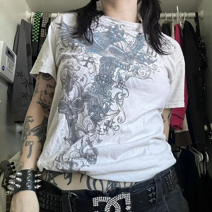 Person with tattoos wearing a Maramalive™ 2000s Retro Grunge T-shirts Women Y2K Cyber Goth Emo Skull Tees E-girl Graphic Print Short Sleeve Tops Vintage Streetwear and a black belt with a rhinestone buckle, standing in front of a clothes rack.