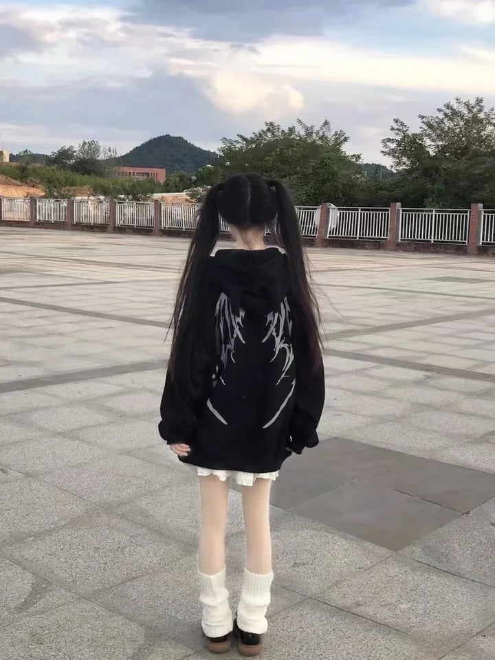 A person with long black hair in pigtails, wearing a trendy Maramalive™ Gothic Zip Up Hoodies Women Mall Goth Tops Streetwear Kawaii Hooded Sweatshirt 2022 Autumn Pullovers with a white wing design on the back, stands in an open paved area with mountains and trees in the background. The outfit embodies the perfect blend of streetwear and women's fashion.