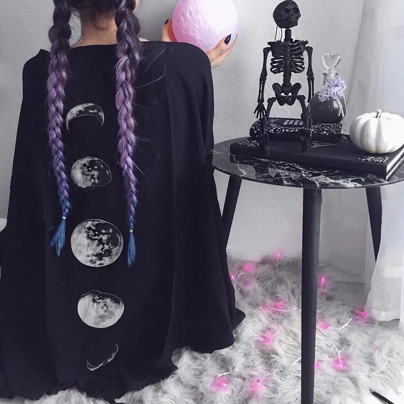 A person with long, braided purple hair sits on the floor facing away, wearing a Maramalive™ Gothic Black Retro Moon Print Tassel Cloak Female Autumn and Winter Dark V-neck Loose Top Bat Cloak Goth Jacket Women, embodying a gothic style. Nearby, a table holds a skeleton figurine and other intriguing items.