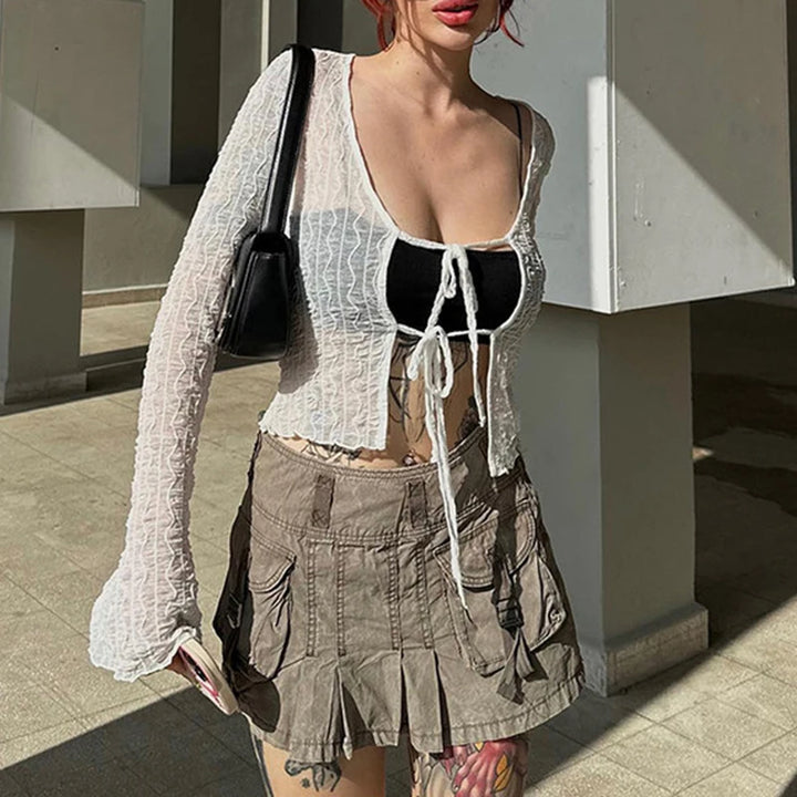 Person wearing a white mesh cardigan over a black top, paired with a beige pleated skirt, standing outdoors. The person is carrying a black shoulder bag, complemented by the slim fit of the ensemble that adds a touch of Gothic fashion to the look. The ensemble features the Maramalive™ Goth Dark Folds Mall Gothic Lace Up Sexy Blouses Grunge Y2k Fairycore Long Sleeve T-shirts Female Transprent Skinny Fashion Tops.