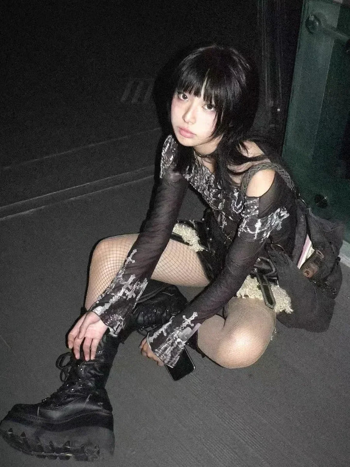 Young person with black hair sits on the floor in a dark streetwear outfit, including fishnet stockings and platform boots, holding a phone in one hand while wearing the Maramalive™ Gothic Goth Black Mesh Tops Women Grunge Aesthetic Off Shoulder Graphic Crop T Shirts See Through Trashy Y2k 2000s Tees.