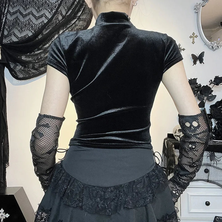 A person in a black velvet dress with lace gloves stands with their back to the camera, in a room decorated with black and white Gothic decor, including black lace fabric, skull accessories, and Maramalive™ Goth Dark Cross Sheer Mall Gothic Women T-shirts Grunge Aesthetic Punk Sexy Emo Black Top Streetwear Fashion Alternative Clothes, epitomizing Gothic style.