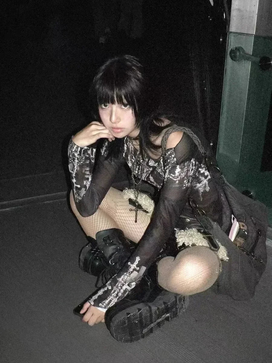 A person with dark hair and bangs sits on the floor, wearing a Maramalive™ Gothic Goth Black Mesh Tops Women Grunge Aesthetic Off Shoulder Graphic Crop T Shirts See Through Trashy Y2k 2000s Tees, fishnet stockings, and black boots, embracing a streetwear style while looking at the camera in a dimly lit room.