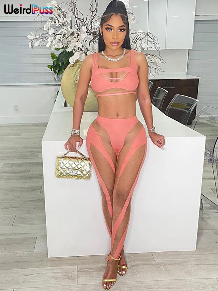 A woman in a sheer, pink outfit with cut-outs and ankle-length pants stands in a modern kitchen, holding a gold handbag. She is wearing gold sandals and jewelry, with white flowers in the background. Perfect for a sexy & club look. She is wearing the Mesh Sexy 2 Piece Set Women Tracksuit Patchwork Fitness Sporty Vest+Leggings Stretch Skinny Midnight Matching Outfits by Maramalive™.