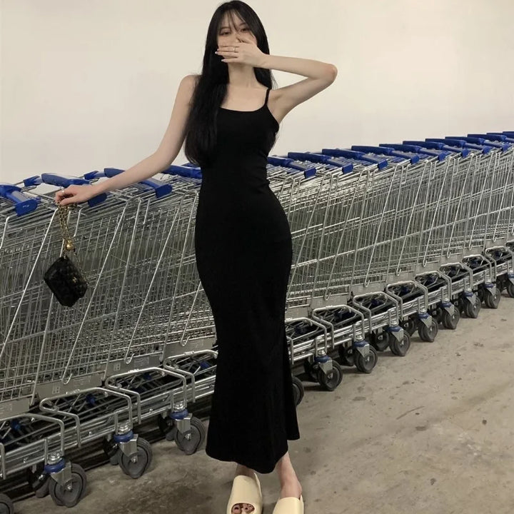 A person in a black Maramalive™ Autumn Pure Sleeveless Dress Women Sexy Sheath Hotsweet Style Fashion All-match Vestido Feminino New Arrival Popular Chic and light-colored footwear stands in front of a row of shopping carts, covering their mouth with one hand and holding a small purse in the other.