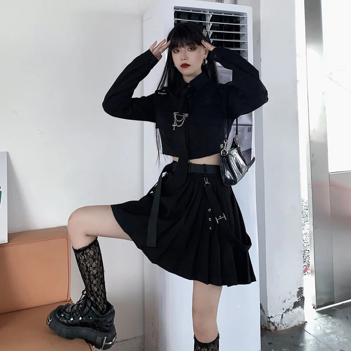 A person dressed in black emo clothing, including a cropped jacket, skirt, and chunky shoes, poses indoors next to an air conditioning unit. They are standing with one foot resting on an orange couch, wearing the EMO Gothic Cargo Shirt Suit Egirl Punk Chain Ribbon Skirts Goth Dress Autumn Streetwear Black Grunge Aesthetic Clothes by Maramalive™.