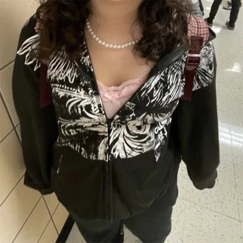 A person with curly hair is wearing a black and white floral jacket with geometric patterns, a pink lace top, and a pearl necklace, standing in a hallway.