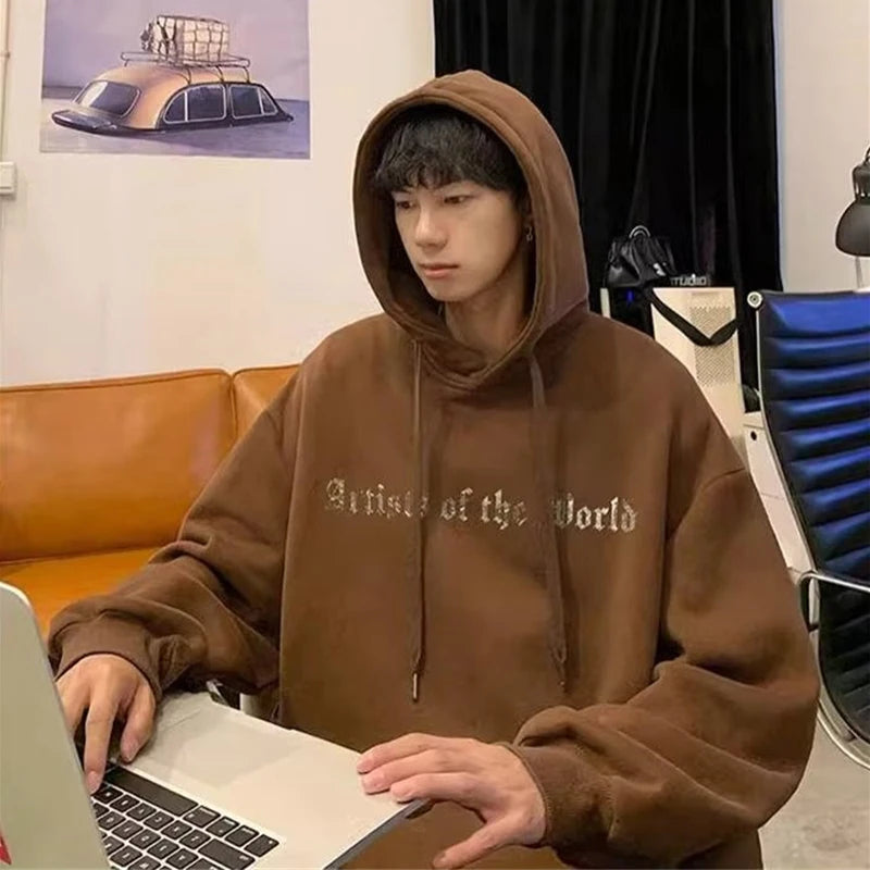 A person in a brown Maramalive™ Autumn Goth Graphic Print Hooded Sweatshirts For Men Oversized Y2K Streetwear Hoodies New In Fashion Pullover Hoodie with "Artist of the World" text works on a laptop at a desk, with a poster of a car in the background.