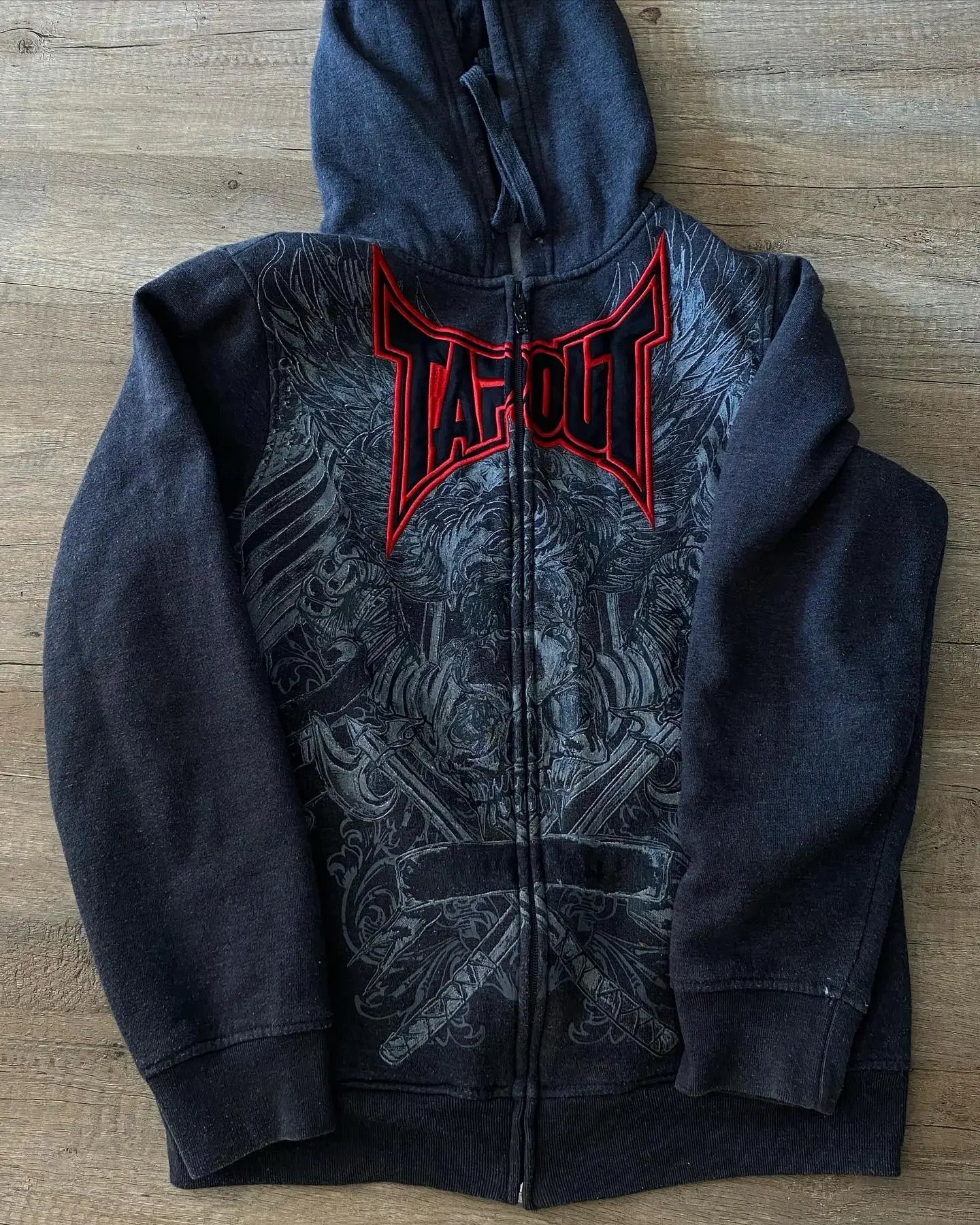A **Gothic Skull Pattern Zip Up Hoodies Men Y2K Embroidery Hip Hop Long Sleeve Loose Hooded Streetwear Sweatshirt Casual Vintage New** with a detailed skull pattern design, featuring the word "Tapout" in red lettering on the front. The sweatshirt, from **Maramalive™**, is laid flat on a wooden surface.