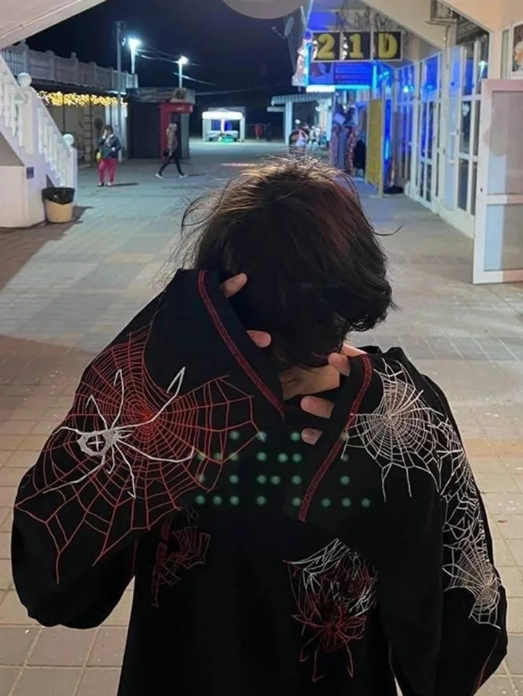 A person with dark hair is seen from behind, wearing a black hoodie with red and white web designs. They are standing on an outdoor walkway at night, with a store behind them lit in blue. The geometric pattern teems with Y2K style vibes, perfect for the Maramalive™ Deeptown Y2k Gothic Spider T Shirt Women Goth Dark Streetwear Design Tees Black Long Sleeve Top 2023 Autumn Spring collections.