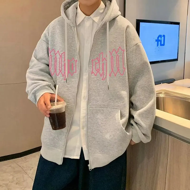 A person in a grey, gothic-style Maramalive™ Autumn Men's Letter Foam Print Zip up Hoodies Y2K Goth Streetwear Loose Sweatshirts Female Hip Hop Oversized Hoodie Tracksuit with pink lettering stands by a counter, holding a cup of iced coffee.