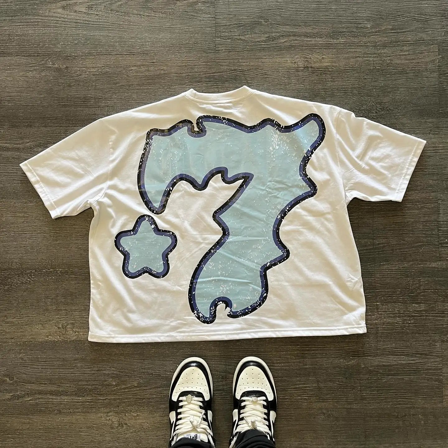 An American 777 graphic t shirts print oversized gothic Smart Casual streetwear graphic y2k tops goth men clothes by Maramalive™ featuring a large, abstract blue graphic design lays flat on a wooden floor, with a pair of sneakers visible at the bottom edge of the image, exuding Harajuku streetwear vibes.