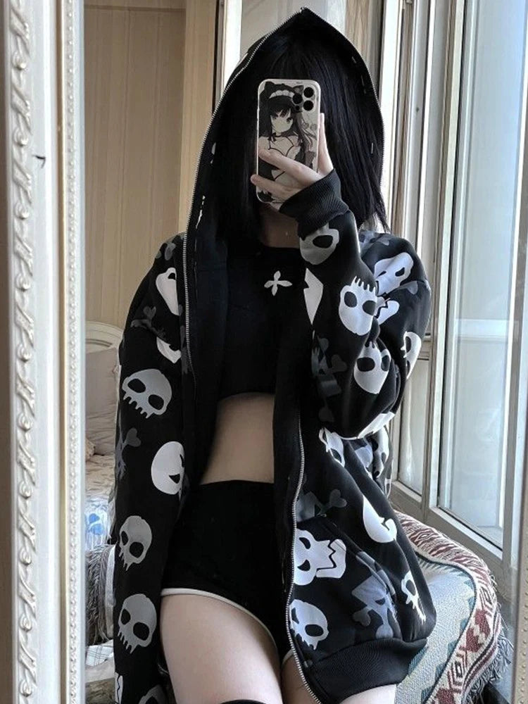 Person in a skull-patterned Maramalive™ Cyberpunk Y2k Sweatshirt Women Mall Goth Skull Printed Long Sleeve Zipper Cardigan Hoodie Emo Alt Indie Clothes taking a mirror selfie with a phone, wearing a black crop top and shorts, and sitting by a window.