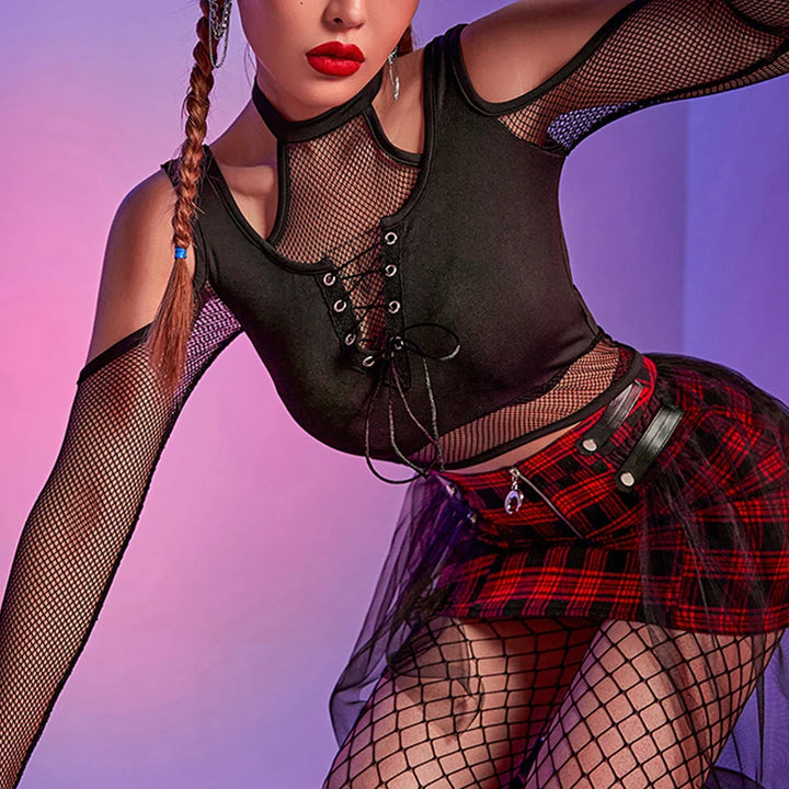 A person dressed in a Goth Dark Techwear Fishnet Open Shoulder Halter T-shirts Mall Gothic Grunge Black Bandage Crop Tops Women Punk Sexy Alt Clothing by Maramalive™, a red plaid skirt with a sheer overlay, and fishnet stockings is posing against a gradient pink and purple background, embodying the dark grunge goth punk aesthetic.