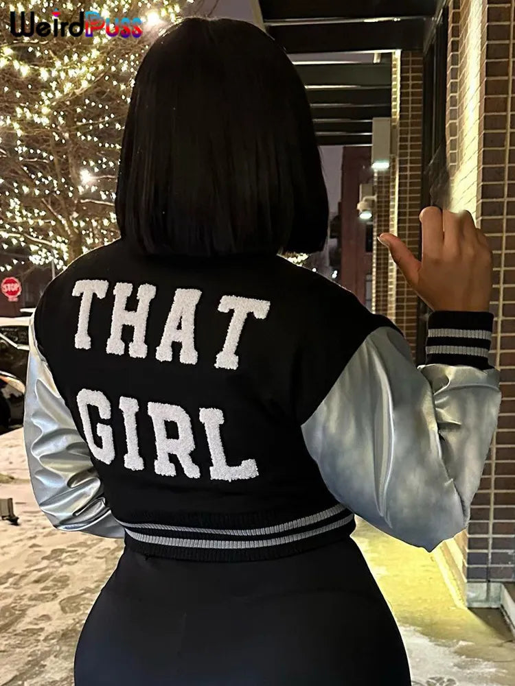 A person with a bob haircut is wearing a black and silver Maramalive™ Fluffy Letter Short Jacket Varsity Women Hipster Leather Sleeve Patchwork Y2K Baseball Casual Wild Streetcoat Outwear that reads "THAT GIRL" on the back, standing outside near a well-lit tree and brick wall at night.
