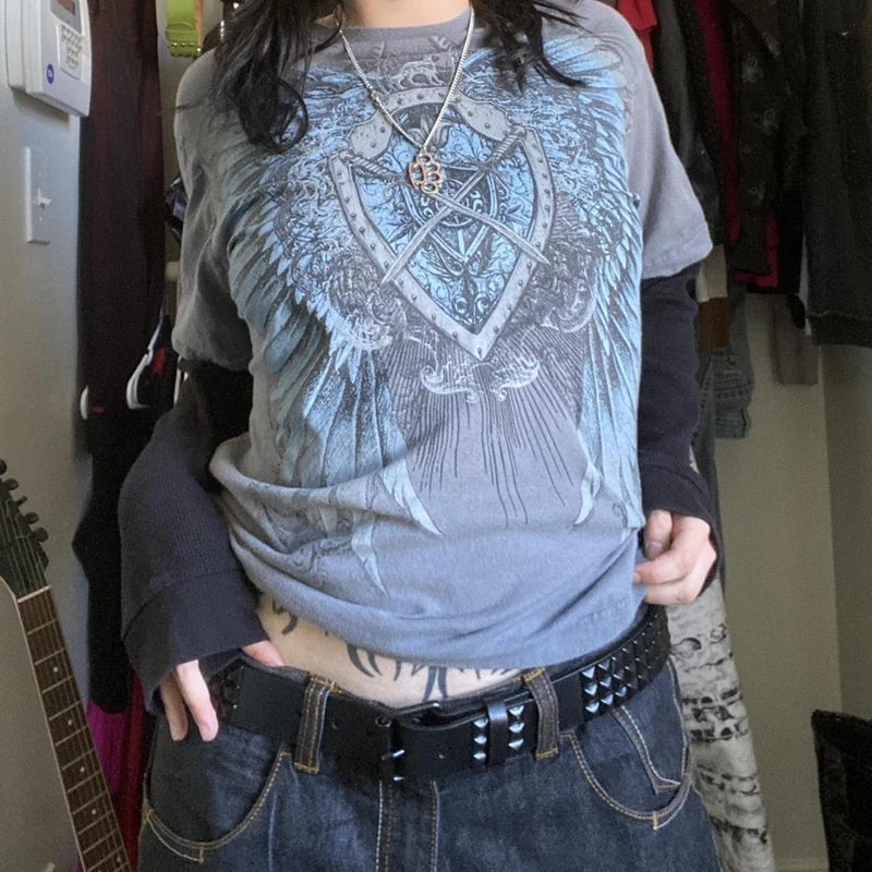 Person wearing a Maramalive™ 2000s Retro Mall Goth Graphic Print Loose Tops Cyber Y2K Grunge Vintage Oversize T-shirt Fake 2 Pice Patchwork Long Sleeve Tees with a shield and wings design, layered over a long-sleeve shirt, black studded belt, and jeans. A guitar and various clothing items are visible in the background.