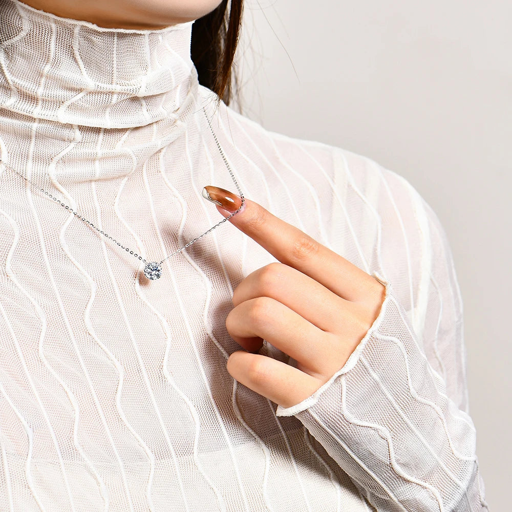 A person wearing a white, sheer textured top points to a small Maramalive™ Moissanite Diamond 6.5mm 1CT Necklace For Woman Pendant 925 Silver Necklace For Women Chains Party Bridal Fine Jewelry with their finger.