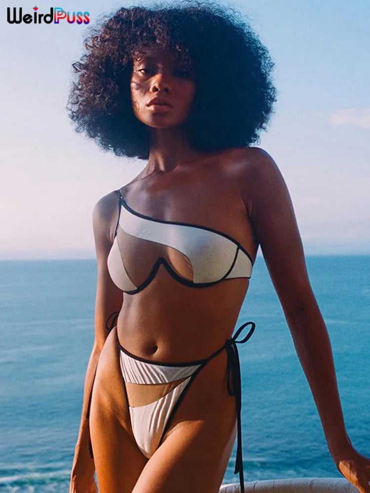 A person with curly hair stands by the ocean wearing a white and black asymmetric bikini made of high stretch fabric, Maramalive™ Beach Style Women Bikini 2 Piece Set Sexy Mesh Patchwork One Shoulder Bra Top+Bandage Briefs Vacation Trend Clubwear, perfect for Summer 2022.