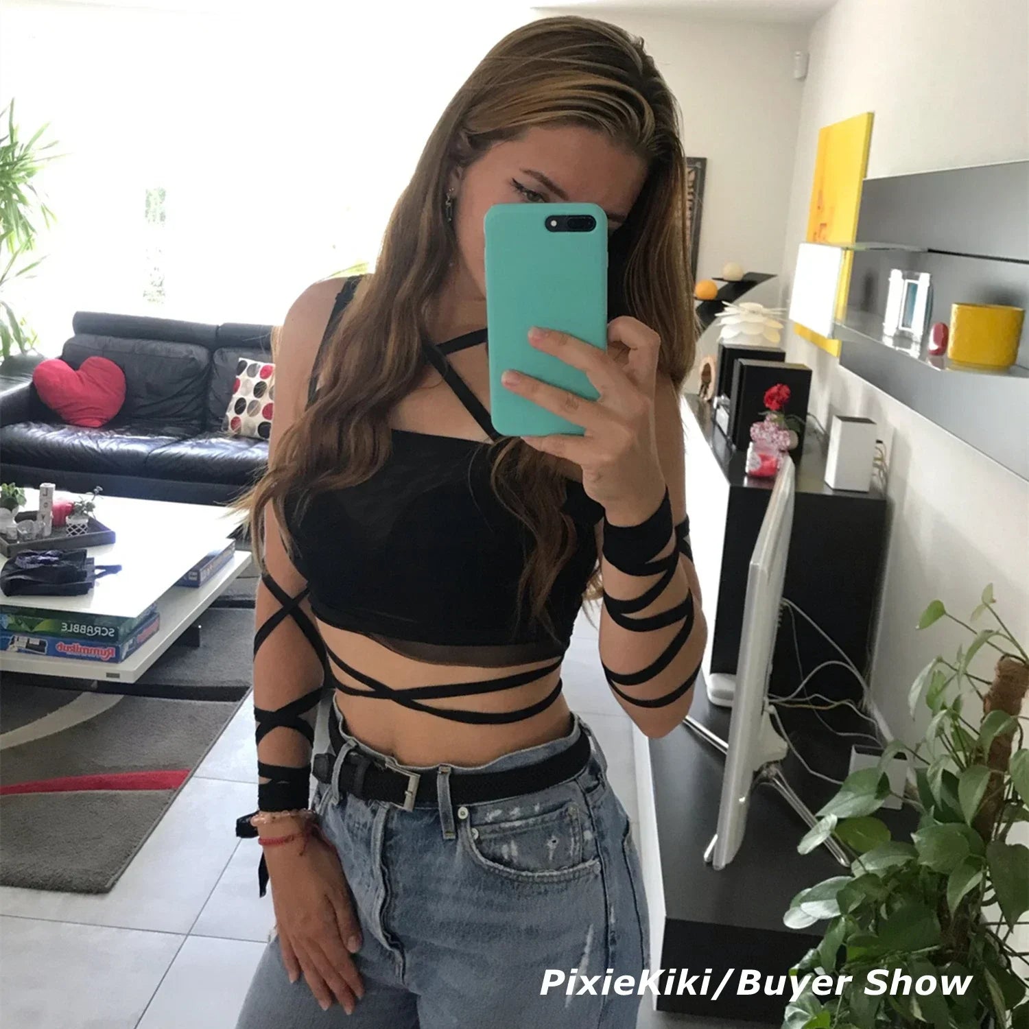 Person taking a mirror selfie in a modern living room. They are wearing a Maramalive™ Black Mesh Lace Up Bandage Crop Top Fairy Grunge Aesthetic Clothes Cyber Y2k Mall Goth Tanks Sexy Clothing P94-BZ14 and blue jeans, holding a phone with a teal case. Furniture and decor are visible in the background.