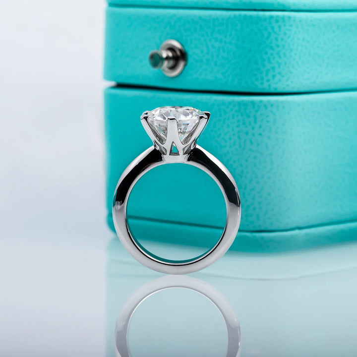 A Maramalive™ Moissanite Solitaire Rings: Stunning Engagement Jewelry is standing upright in front of a blue jewelry box with a metal clasp.