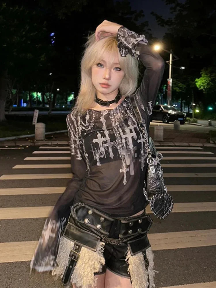 A person with blonde hair wearing a Maramalive™ Gothic Goth Black Mesh Tops Women Grunge Aesthetic Off Shoulder Graphic Crop T Shirts See Through Trashy Y2k 2000s Tees and dark slim fit shorts stands on a street at night, posing with one arm raised and holding a black bag.