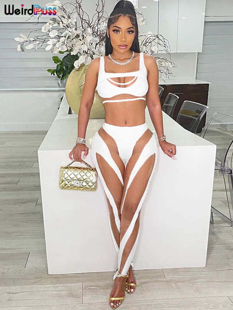A woman in a Maramalive™ Mesh Sexy 2 Piece Set Women Tracksuit Patchwork Fitness Sporty Vest+Leggings Stretch Skinny Midnight Matching Outfits is standing indoors, leaning on a countertop. She is holding a gold purse and wearing sandals. A flower arrangement is visible in the background, adding to the stylish ambiance.