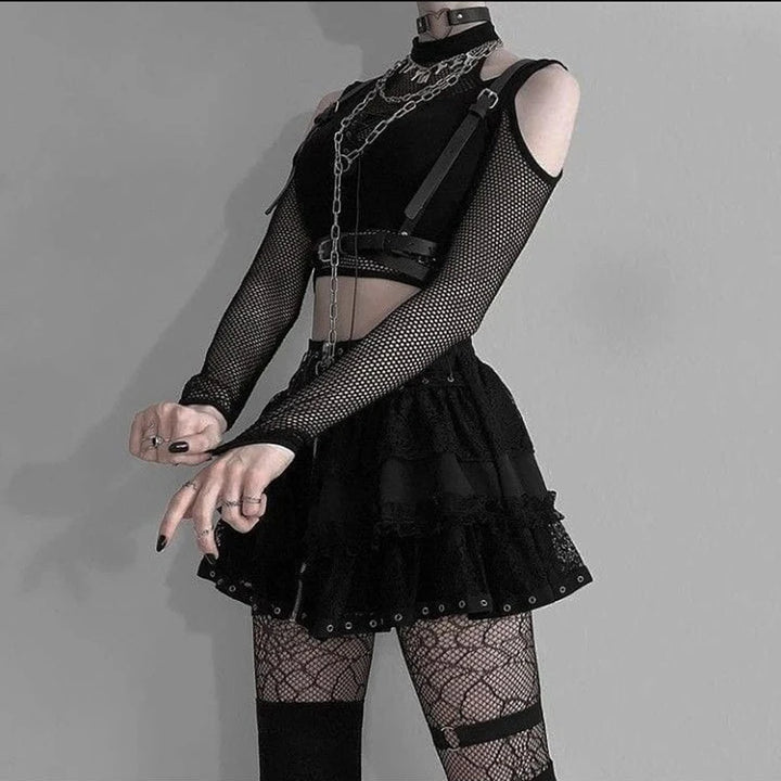 Person clad in a grunge goth punk look, featuring fishnet patchwork sleeves, a choker, chains, a Goth Dark Techwear Fishnet Open Shoulder Halter T-shirts Mall Gothic Grunge Black Bandage Crop Tops Women Punk Sexy Alt Clothing from Maramalive™, a layered skirt, thigh-high stockings, and black painted nails. They stand against a plain gray background.