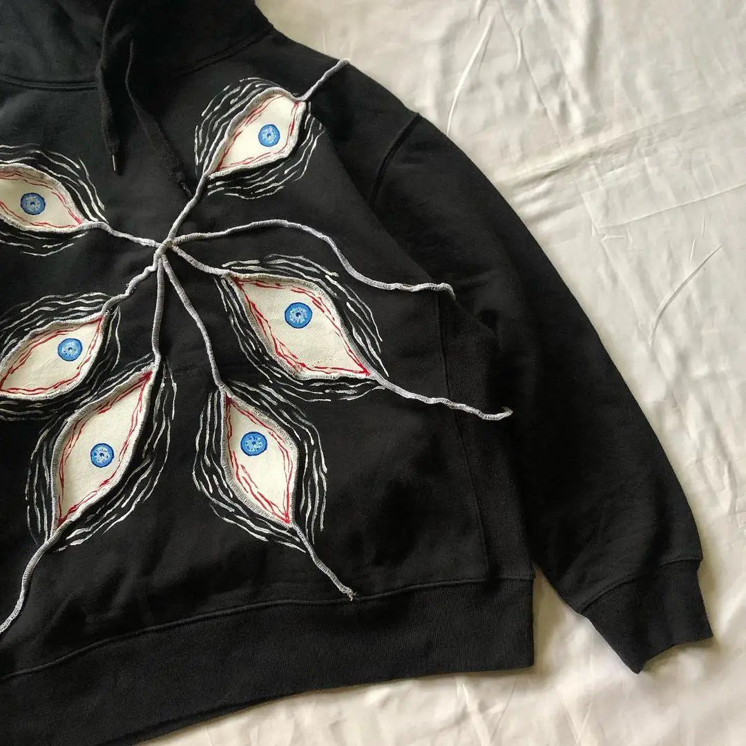 A Maramalive™ 2024 Fashion Goth Eye Mask Black Hooded Sweatshirt Retro Y2K Hip Hop Street Punk Casual Loose Jacket for Men and Women with abstract eye designs featuring blue irises and red veins is laid out on a white surface. White strings extend from the center of the design, making it perfect for Autumn/Winter casual pullovers in a women's loose fit.