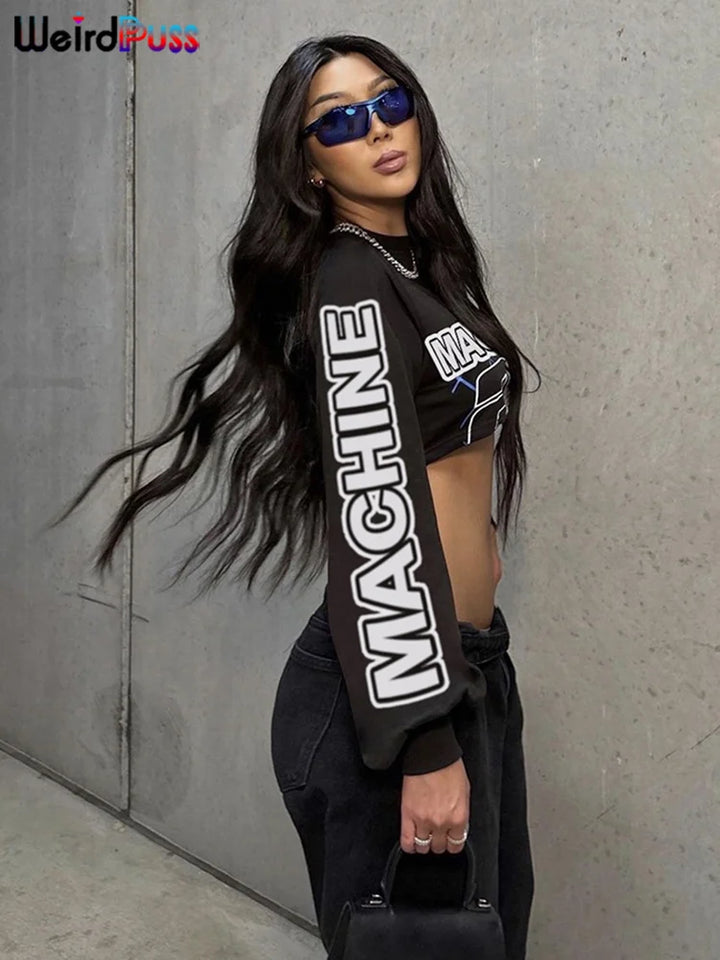 A person with long hair wears sunglasses, the Maramalive™ Letter Hipster 2 Piece Set Women Sporty Casual Skinny Lantern Sleeve Crop Tops+Skirts Trend Streetwear Matching Suits with "MACHINE" on the sleeve, and a black purse in hand, posing against a plain background.