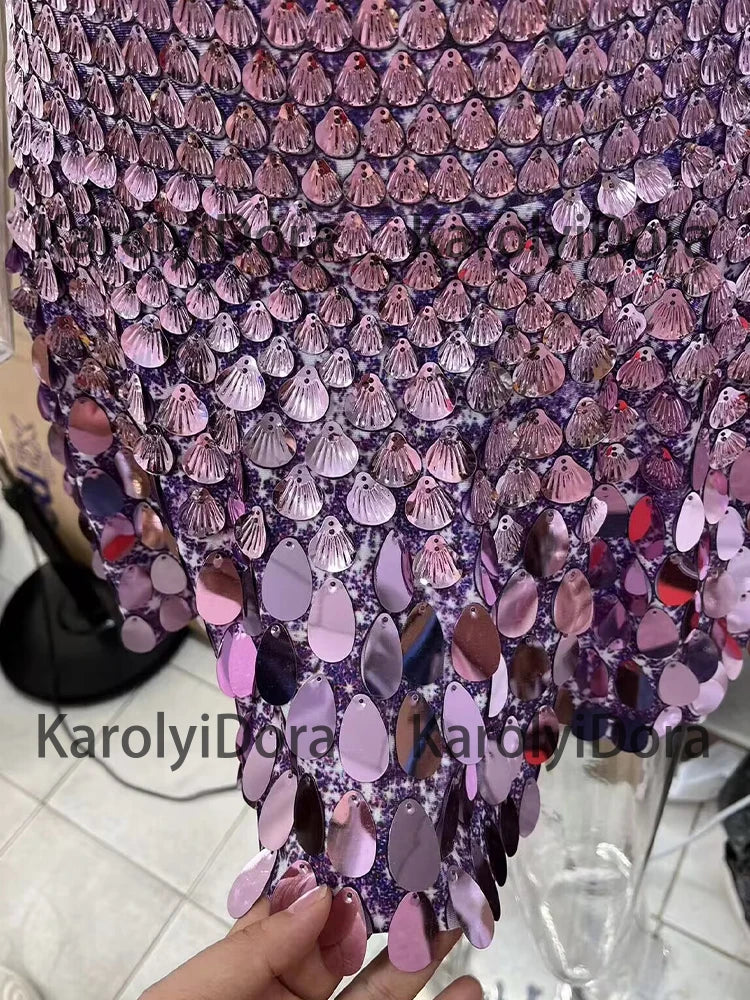A close-up of a hand holding a fabric adorned with shiny, purple, and pink sequins in various shapes, reminiscent of the intricate designs seen on Maramalive™ Sexy Black Shell Sequins Mini Dress Nightclub Dance High Quality Performance Clothing Bodysuit Birthday Red Dresses worn in Chinese Folk Dance by women.
