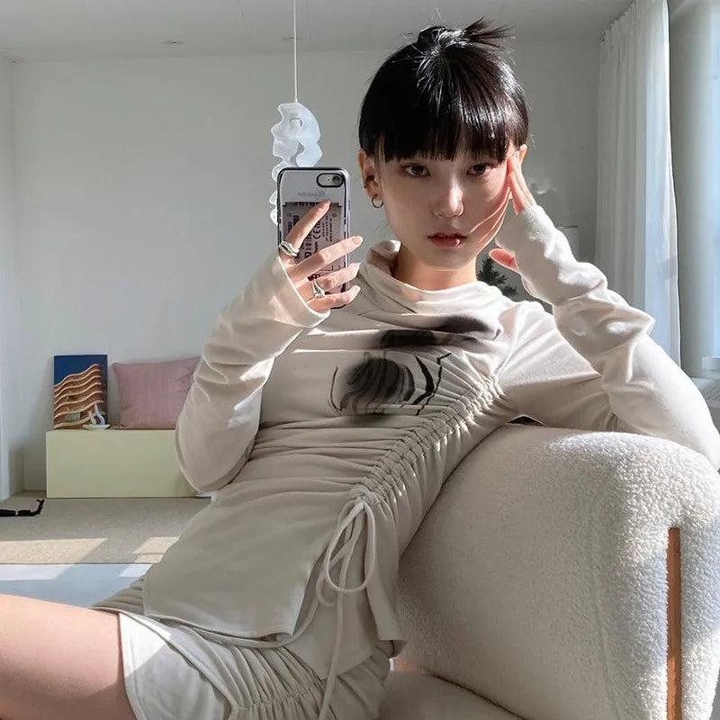 A person with short dark hair, wearing a light-colored Grunge Irregular Tops Dark Aesthetics Bandage T-shirt Mall Goth Y2k Tight Tops Alternative Streetwear Fashion Emo by Maramalive™, takes a mirror selfie while sitting on a white chair in a brightly lit room.