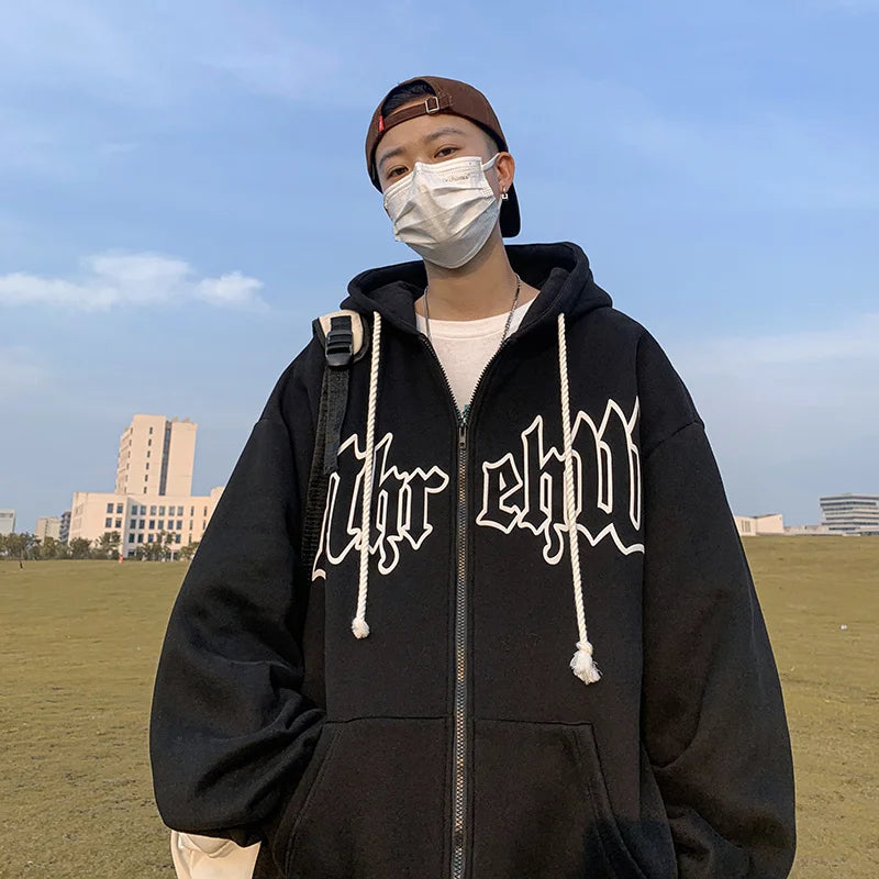 A person wearing a black Maramalive™ Autumn Men's Letter Foam Print Zip up Hoodies Y2K Goth Streetwear Loose Sweatshirts Female Hip Hop Oversized Hoodie Tracksuit with large white text on the front, a white face mask, and a baseball cap stands outdoors on a grassy field with buildings in the background.