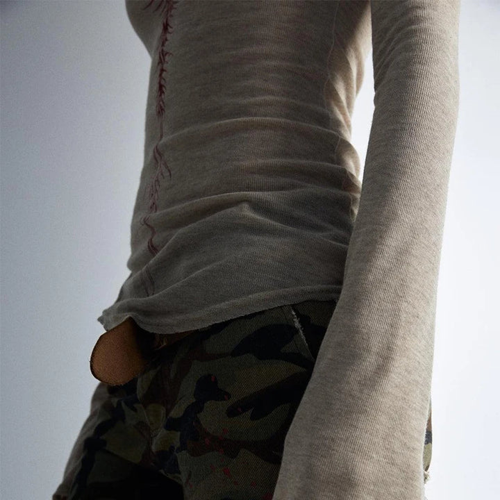 Close-up of a person wearing a fitted, long-sleeve beige top with an elongated sleeve, and camouflage pants. The image, centered on the garments' textures and colors, showcases elements of vintage style in its elegant simplicity. They are wearing the 90s Vintage E-girl Emo Clothes Fires Graphic Print Y2K Grunge T-shirt Women Retro Long Sleeve Tee Goth Punk Streetwear by Maramalive™.