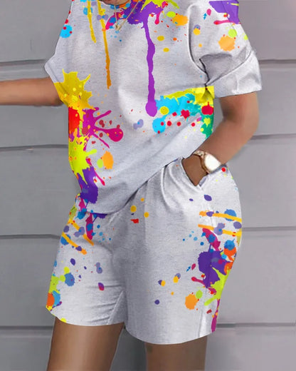 Person wearing a grey, paint-splattered outfit from Maramalive™ with Plus Size Matching Sets Women Graffiti Tie Dye Shirts Tops And Short Pants Female Fashion Tracksuits 2023 Summer Two Piece Sets, both featuring colorful splashes. Embracing casual style, the person is leaning against a grey wall with one hand in their pocket.