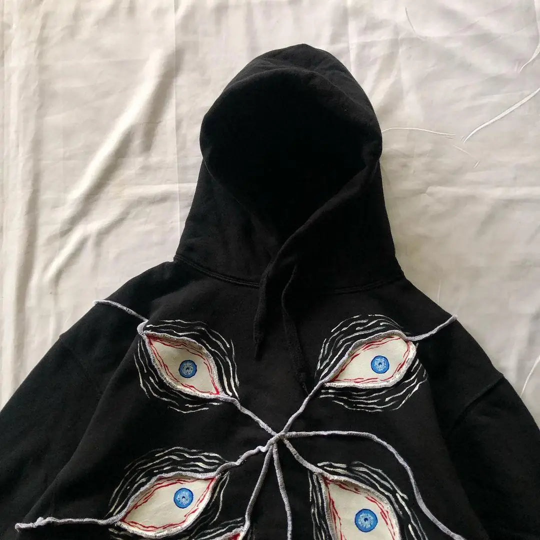 A Maramalive™ 2024 Fashion Goth Eye Mask Black Hooded Sweatshirt Retro Y2K Hip Hop Street Punk Casual Loose Jacket for Men and Women with an abstract design featuring four intricate eyes in various colors, connected by loose, white threads, lies on a white fabric backdrop. Perfect for Autumn/Winter casual pullovers and women's loose fit styles.