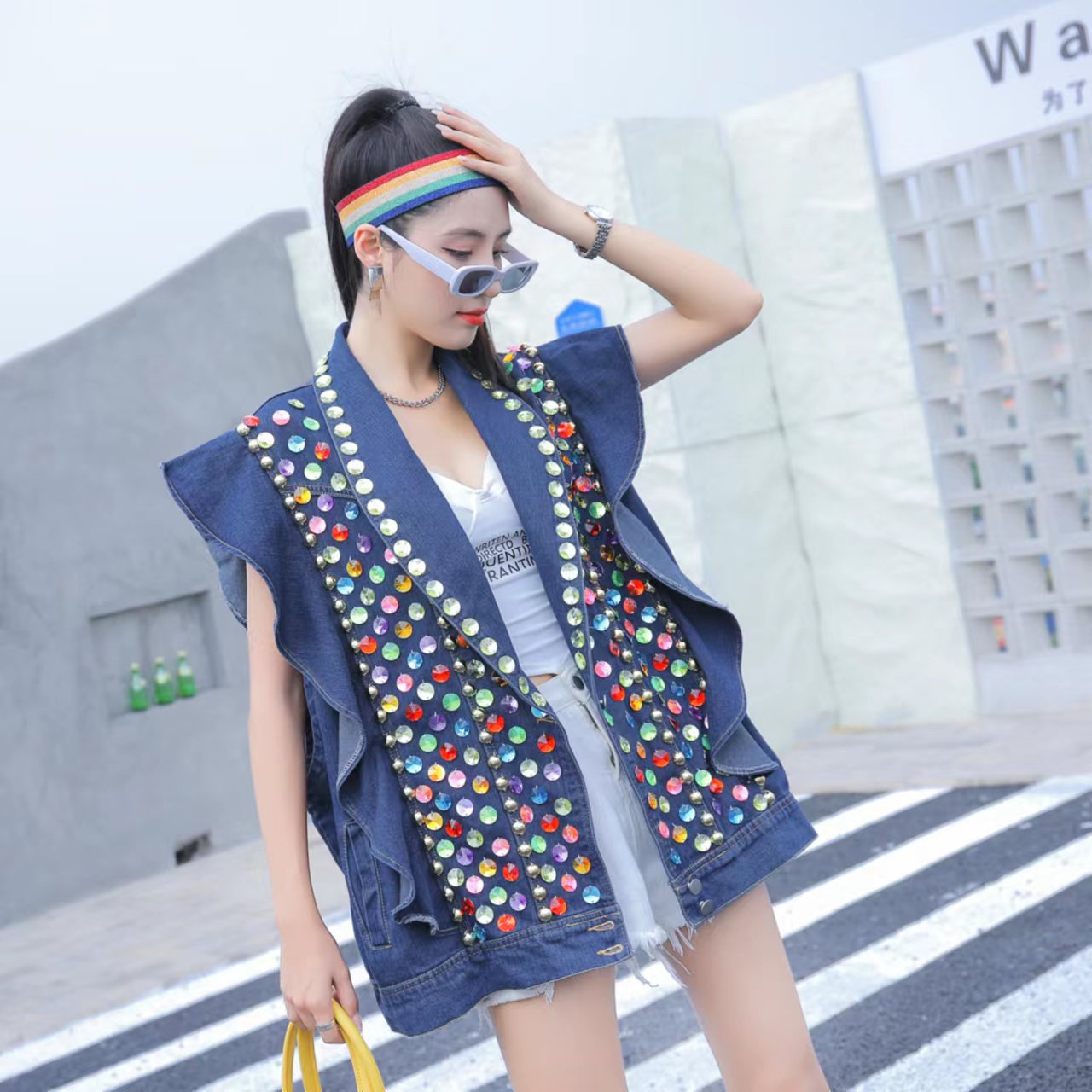 A woman wearing a Maramalive™ Heavy Duty Diamond Studded Denim Vest With Wooden Ear Edge, white shorts, sunglasses, a rainbow headband, and holding a yellow bag, stands on a crosswalk touching her head.