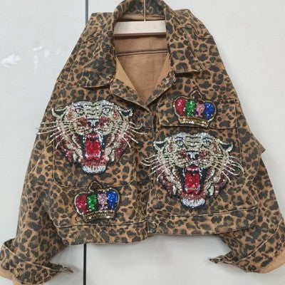 A Leopard Print Studded With Nails Bead Mesh Gauze Stitching Denim Short Coat Female by Maramalive™, crafted from triacetate fiber, boasts two embroidered tiger heads with open mouths and crowns above them on the front pockets, hanging elegantly on a hanger.