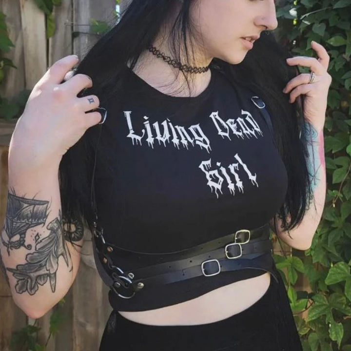A woman with long black hair wears a "Living Dead Girl" shirt, black choker, and strappy belt. She has tattoos on her arm and hand, and greenery is visible in the background. Her outfit, featuring a Maramalive™ Gothic Style Printed Top Short Sleeve, showcases edgy fashion sense with a hint of rebellion.