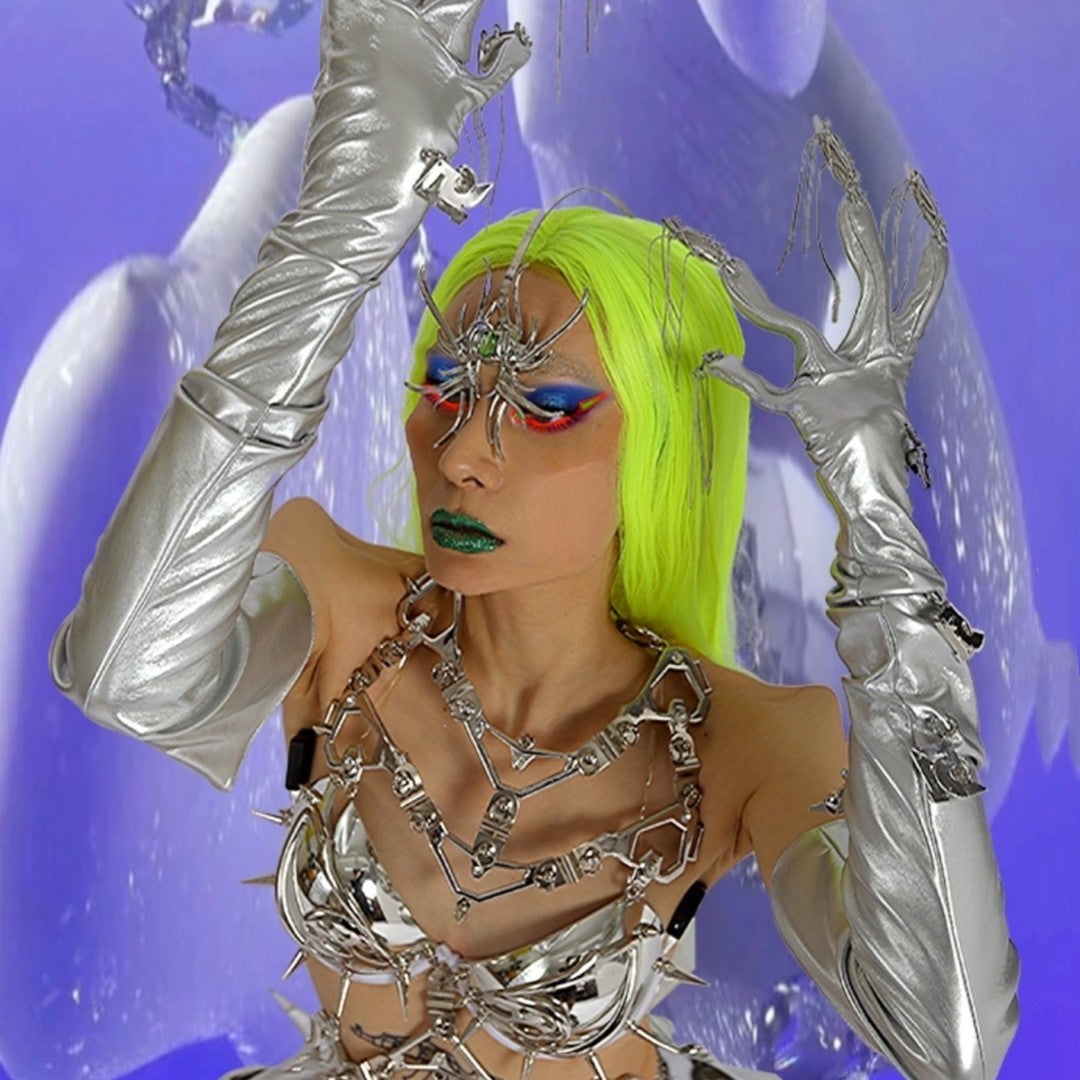 Person with neon green hair, wearing dramatic makeup, silver gloves, and futuristic spiked accessories, adorned with striking electroplated jewelry and facial decoration, posed against a blue abstract background in a Punk Futuristic Silver Metallic Performance Suit by Maramalive™.