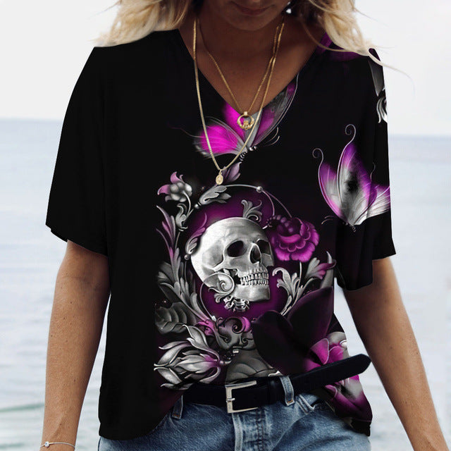 A woman wearing a Maramalive™ Ladies' Printed V-Neck Tee | Chic Women's Graphic Tees with a vivid design featuring a skull, purple flowers, and butterflies. The flattering fit accentuates her figure as she pairs it with a pendant necklace and belt, standing against a blurred outdoor background.